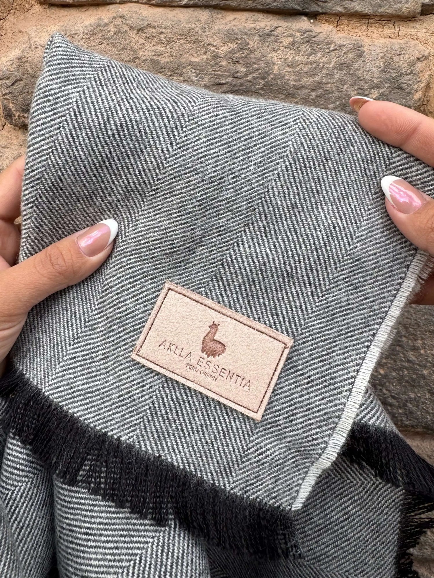 Mano sosteniendo una manta de baby alpaca gris con etiqueta Aklla Essentia, símbolo del lujo sostenible hecho en Perú.